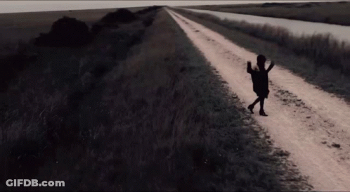 A Woman Walking Down A Dirt Road With A Purse In Her Hand GIF