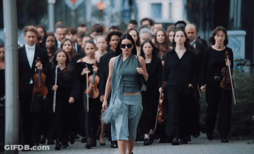 A Woman Walking In Front Of A Large Group Of People Holding Violins GIF