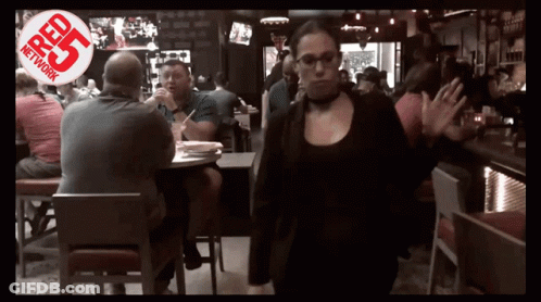 A Woman Walking Through A Crowded Restaurant With A Red R25 Network Logo Behind Her GIF