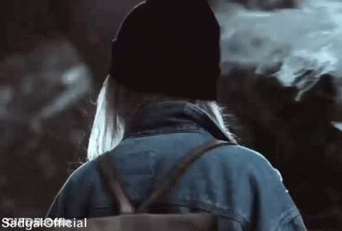 A Woman Wearing A Beanie And A Denim Jacket Is Walking With A Backpack .. GIF