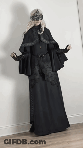 A Woman Wearing A Black Cape And A Mask Stands In Front Of A White Wall GIF