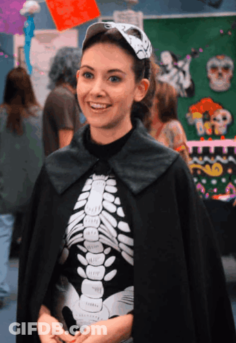 A Woman Wearing A Black Cape And A Skeleton Shirt Smiles GIF