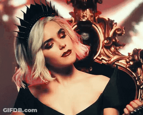 A Woman Wearing A Black Crown And A Black Dress Is Sitting On A Chair . GIF