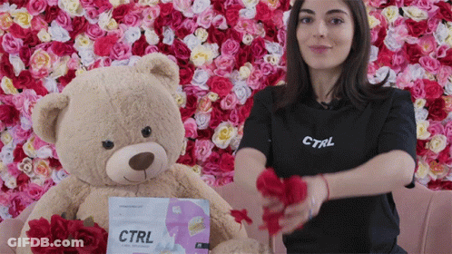 A Woman Wearing A Black Ctrl Shirt Holds A Teddy Bear In Front Of A Wall Of Flowers GIF