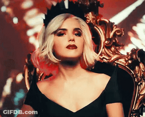 A Woman Wearing A Black Dress And A Crown Is Sitting On A Chair . GIF