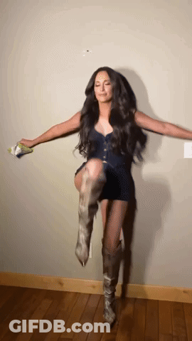 A Woman Wearing A Black Dress And Cowboy Boots Is Standing On One Leg GIF