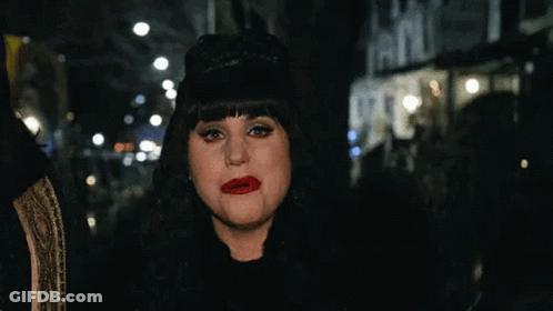 A Woman Wearing A Black Hat And Red Lipstick Is Standing On A City Street At Night . GIF