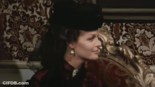 A Woman Wearing A Black Hat Is Sitting On A Bed GIF