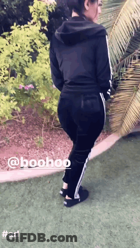 A Woman Wearing A Black Hoodie And Black Pants Is Standing On A Grassy Field GIF