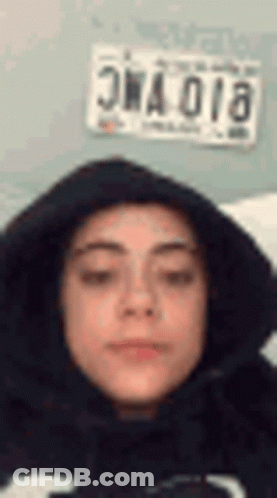 A Woman Wearing A Black Hoodie Is Making A Funny Face In Front Of A License Plate . GIF