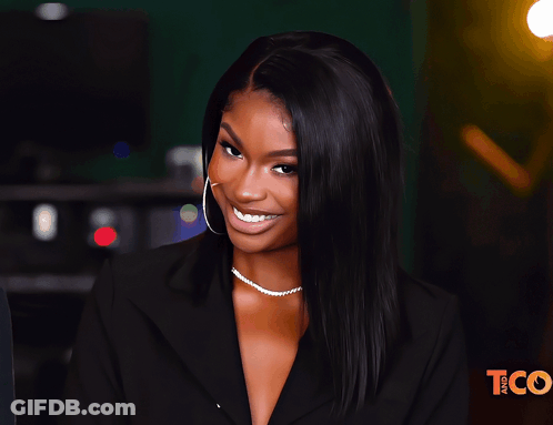 A Woman Wearing A Black Jacket And A Pearl Necklace Smiles In Front Of A Tco Logo GIF