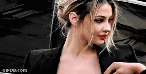 A Woman Wearing A Black Jacket And Red Lipstick Is Standing In Front Of A Black Car . GIF