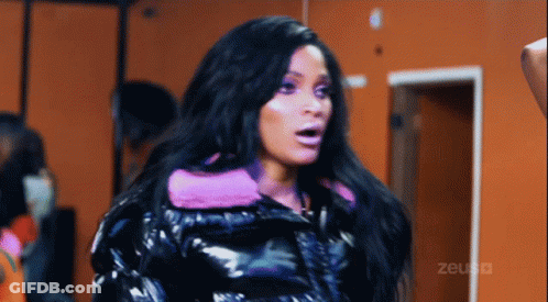 A Woman Wearing A Black Jacket With A Pink Hood Is Standing In Front Of An Orange Wall With The Word Zeus Written On It GIF
