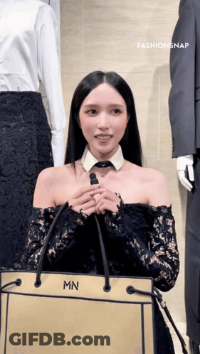 A Woman Wearing A Black Lace Off The Shoulder Top Is Holding A Bag With The Letter Mn On It GIF