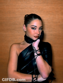 A Woman Wearing A Black Leather Dress And Gloves Is Standing In Front Of A Wooden Wall GIF