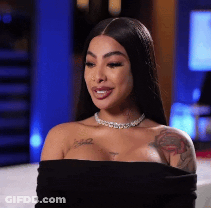 A Woman Wearing A Black Off The Shoulder Top And A Necklace Is Sitting On A Bed . GIF