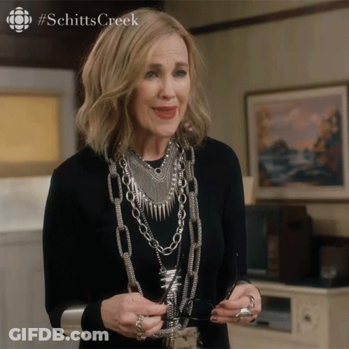 A Woman Wearing A Black Shirt And Silver Jewelry Is Holding A Pair Of Glasses With The Hashtag Schitts Creek On The Top GIF