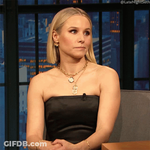 A Woman Wearing A Black Strapless Dress And A Gold Necklace Is Sitting In Front Of A Window GIF
