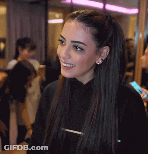 A Woman Wearing A Black Sweater With The Letter T On The Front Smiles GIF