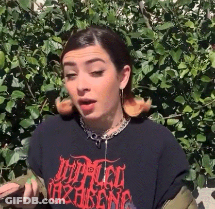 A Woman Wearing A Black T-Shirt And A Necklace Is Standing In Front Of A Tree . GIF