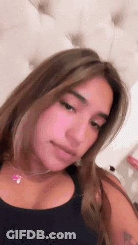 A Woman Wearing A Black Tank Top And A Pink Necklace Is Sitting On A Bed . GIF