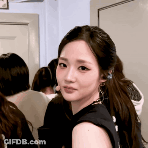 A Woman Wearing A Black Tank Top And Earrings Looks Over Her Shoulder GIF
