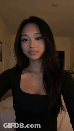 A Woman Wearing A Black Top And A Necklace Is Standing In A Dark Room . GIF