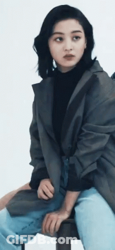 A Woman Wearing A Black Turtleneck And A Grey Jacket Sits On A Stool GIF