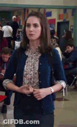 A Woman Wearing A Blue Cardigan And A Floral Shirt Is Standing In A Hallway . GIF
