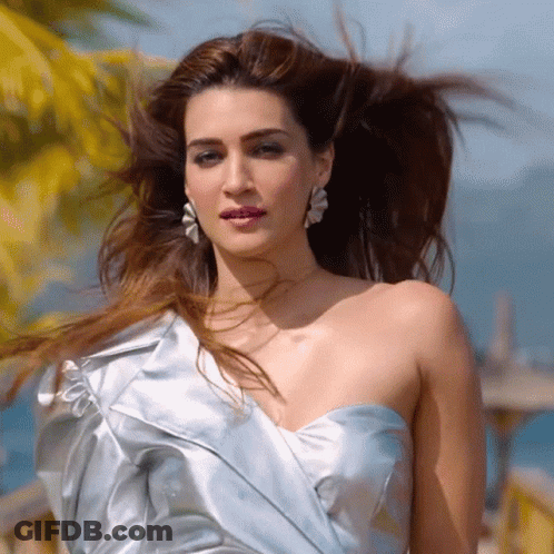 A Woman Wearing A Blue Dress And Earrings Stands In Front Of A Palm Tree GIF