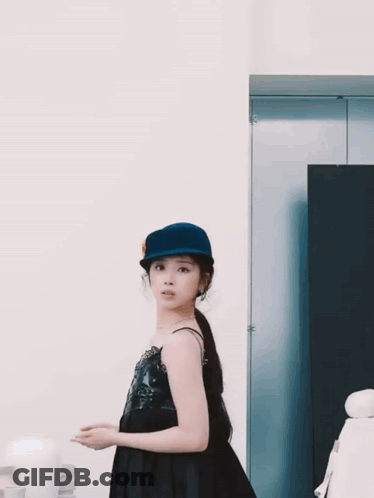 A Woman Wearing A Blue Hat And A Black Dress Looks At The Camera GIF