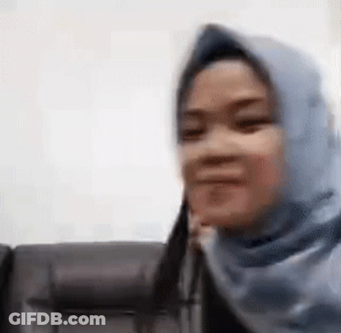 A Woman Wearing A Blue Hijab Is Sitting On A Couch And Smiling . GIF