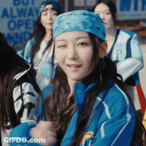 A Woman Wearing A Blue Jacket And A Blue Bandana Is Smiling In Front Of A Sign That Says But Always Open GIF