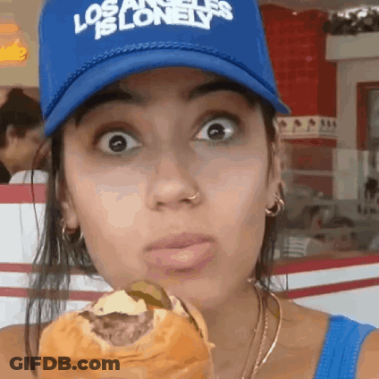 A Woman Wearing A Blue Los Angeles Is Lonely Hat Is Eating A Hamburger . GIF