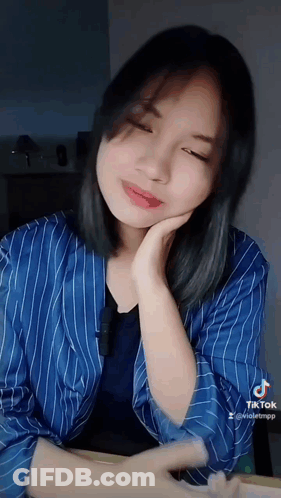 A Woman Wearing A Blue Striped Shirt Is Sitting At A Table With Her Hand On Her Chin . GIF