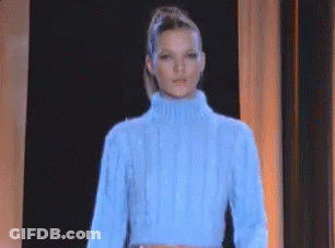 A Woman Wearing A Blue Sweater Is Standing In Front Of A Black Curtain . GIF
