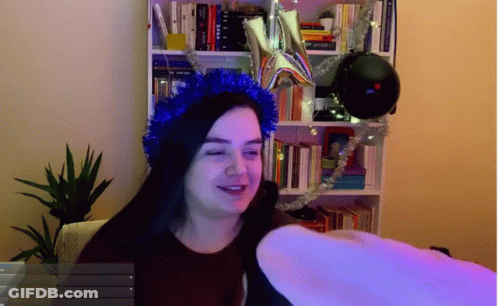 A Woman Wearing A Blue Tinsel Headband Stands In Front Of A Bookcase GIF