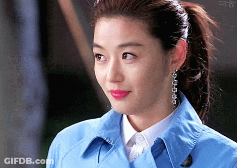 A Woman Wearing A Blue Trench Coat And Earrings Looks At The Camera GIF