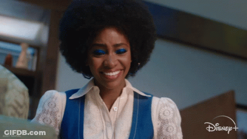A Woman Wearing A Blue Vest And White Shirt Is Smiling For Disney + GIF