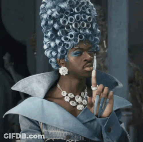 A Woman Wearing A Blue Wig And A Necklace Holds Her Finger Up GIF