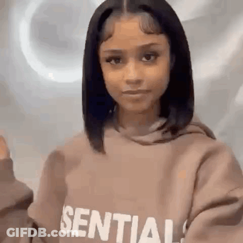 A Woman Wearing A Brown Hoodie Is Standing In Front Of A White Wall . GIF