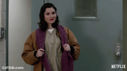 A Woman Wearing A Brown Jacket And A Purple Scarf Stands In Front Of A Netflix Logo GIF