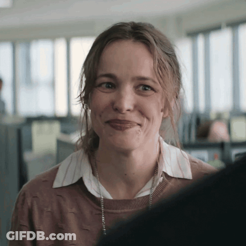 A Woman Wearing A Brown Sweater And A White Shirt Smiles For The Camera GIF