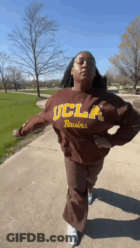 A Woman Wearing A Brown Ucla Bruins Sweatshirt Is Walking Down A Sidewalk . GIF