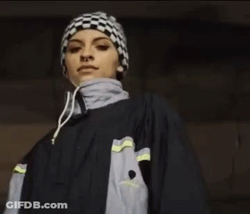 A Woman Wearing A Checkered Hat And A Jacket Is Pointing At The Camera . GIF