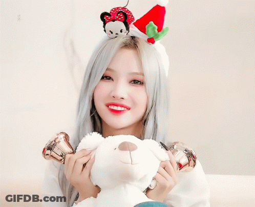 A Woman Wearing A Christmas Hat Holds A Bell In Front Of Her Ear While Holding A Teddy Bear GIF