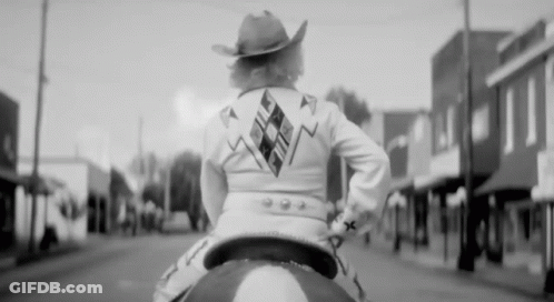 A Woman Wearing A Cowboy Hat Is Riding A Horse Down A Street GIF