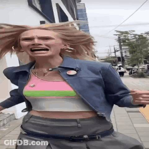 A Woman Wearing A Crop Top And A Denim Jacket Is Running Down A Street GIF