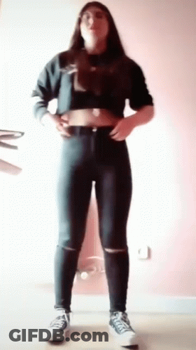 A Woman Wearing A Crop Top And Jeans Is Standing In A Room . GIF