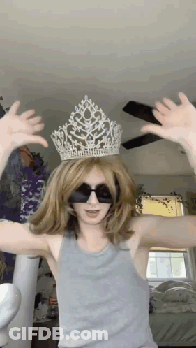 A Woman Wearing A Crown And Sunglasses Holds Her Hands Up In The Air GIF